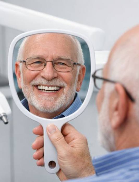 Senior dental patient smiling at his reflection in hand mirror