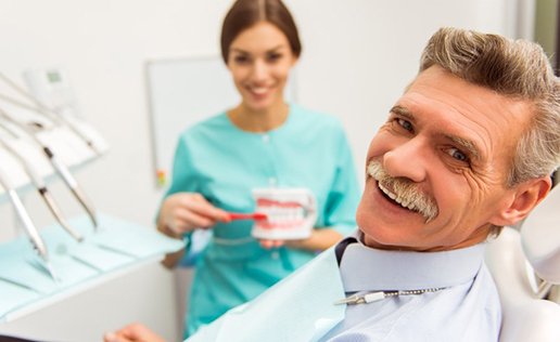 Man smiling to reveal missing teeth
Man in patient’s chair smiling while dentist holds a model denture

